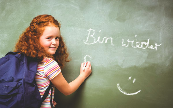 ein Mädchen mit einem Rucksack, das "Bin wieder da! :-)" an eine Tafel schreibt